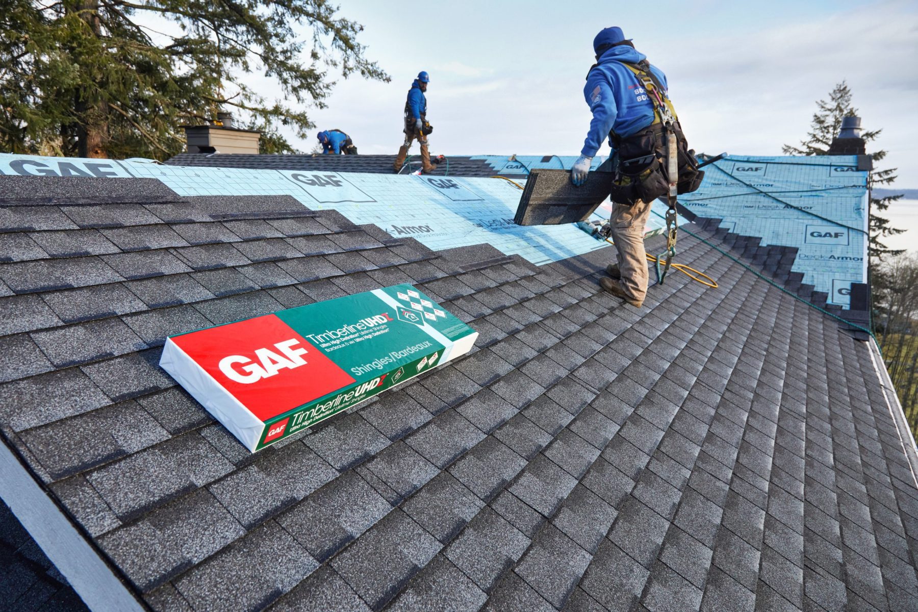 Three employees work on installing new shingles to a roof with a GAF product in the forefront and scenic backdrop